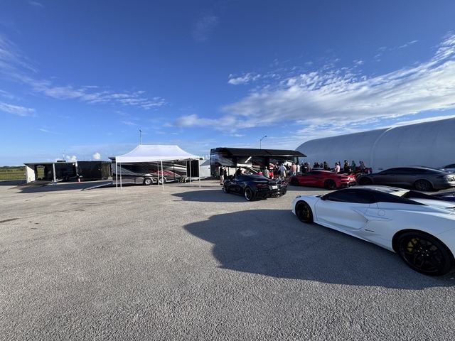 A white sports car in the lot beside transport trailers and the event tent.