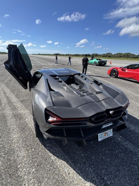 A gray supercar with gullwing doors open, other cars and spectators behind.