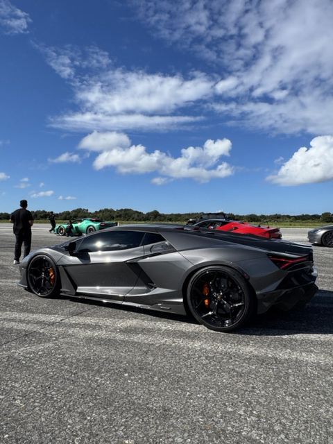 A silver supercar in front of red and turquoise cars on the runway.